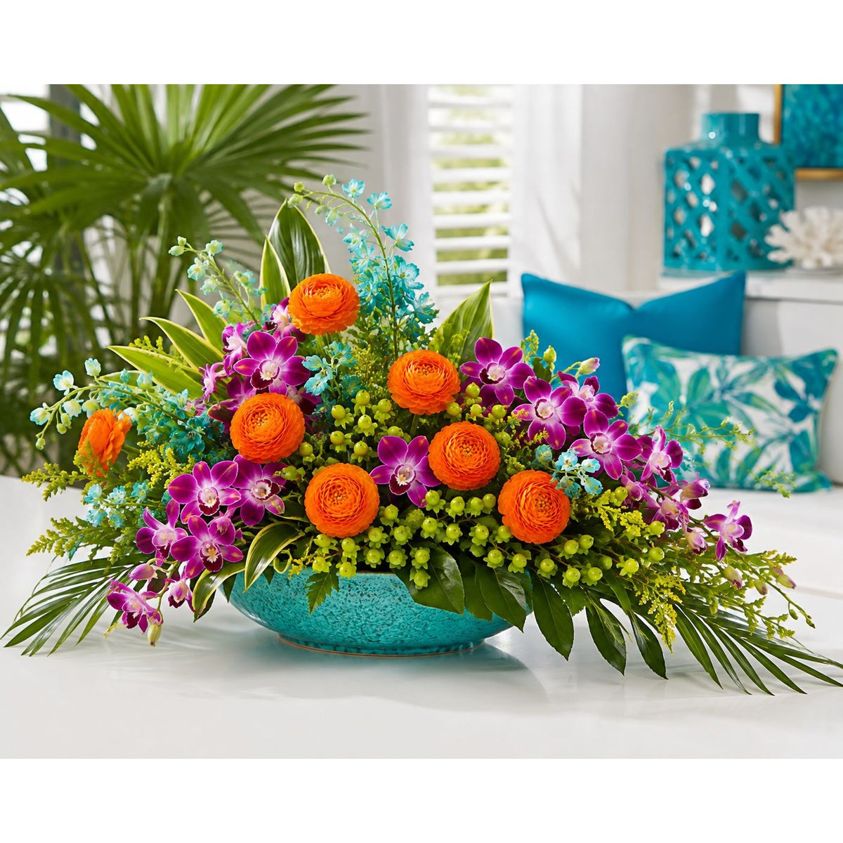A vibrant tropical floral arrangement in a turquoise bowl, featuring bright orange ranunculus, purple orchids, and green foliage, set on a white surface with a blurred turquoise and white tropical background.