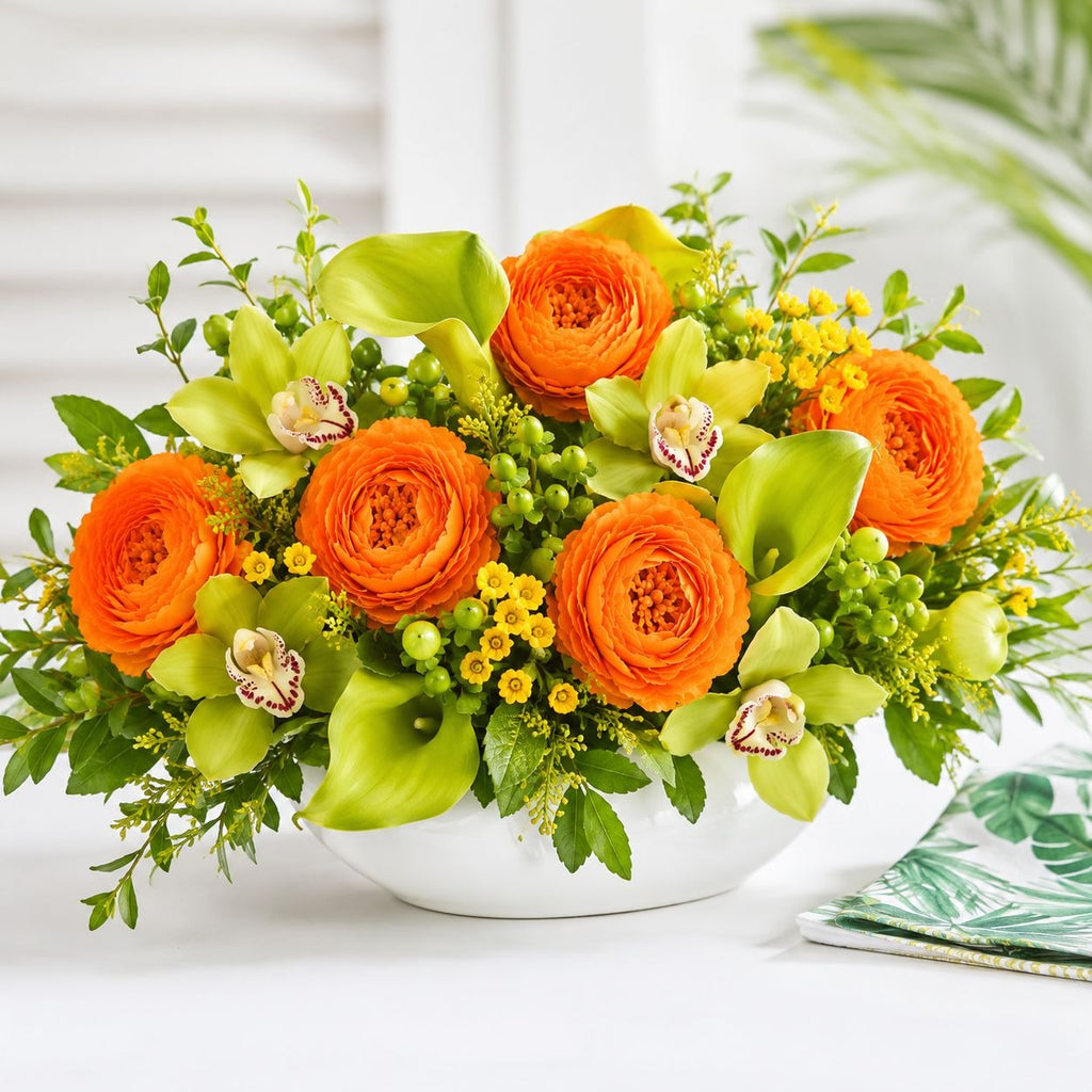 A vibrant floral arrangement featuring bright orange ranunculus, lime green calla lilies, orchids, and small yellow blossoms in a white ceramic bowl, set against a bright, softly blurred background.