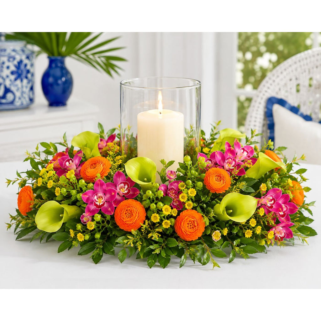 A vibrant floral centerpiece with a lit white pillar candle in a clear glass holder, surrounded by bright orange ranunculus, pink orchids, green calla lilies, and yellow filler flowers on a white table.