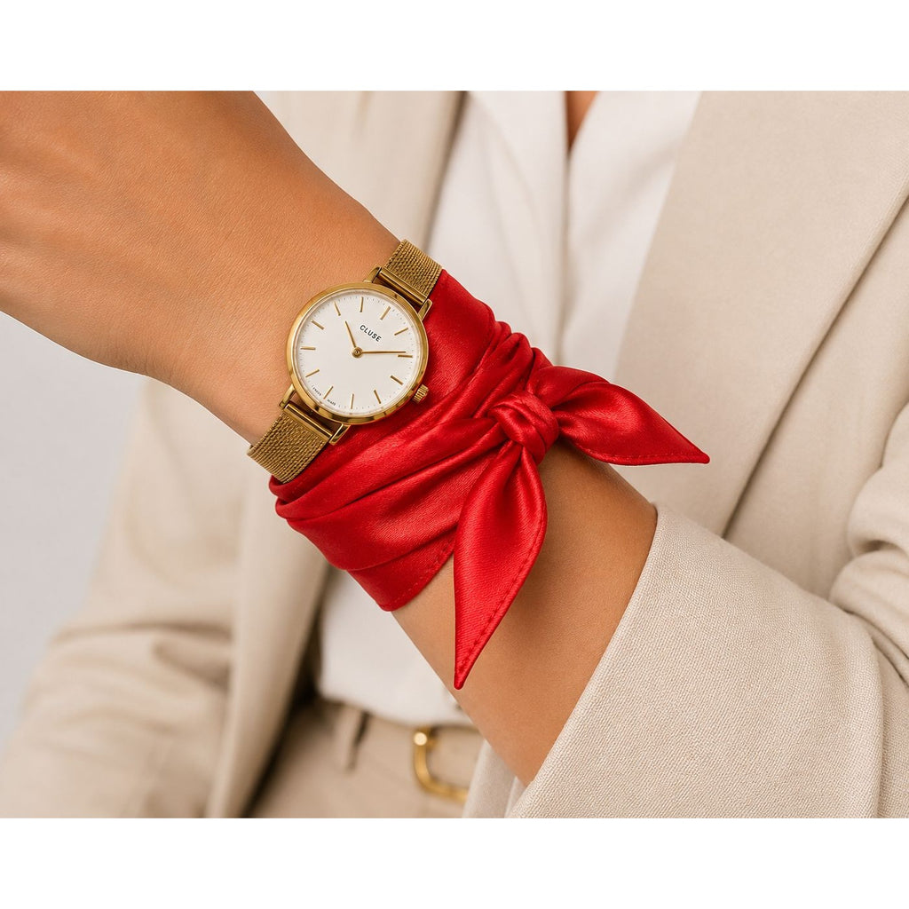 A close-up of a woman's wrist wearing a gold-tone mesh watch with a white dial and a red satin scarf tied around her wrist, against a light beige blazer background.