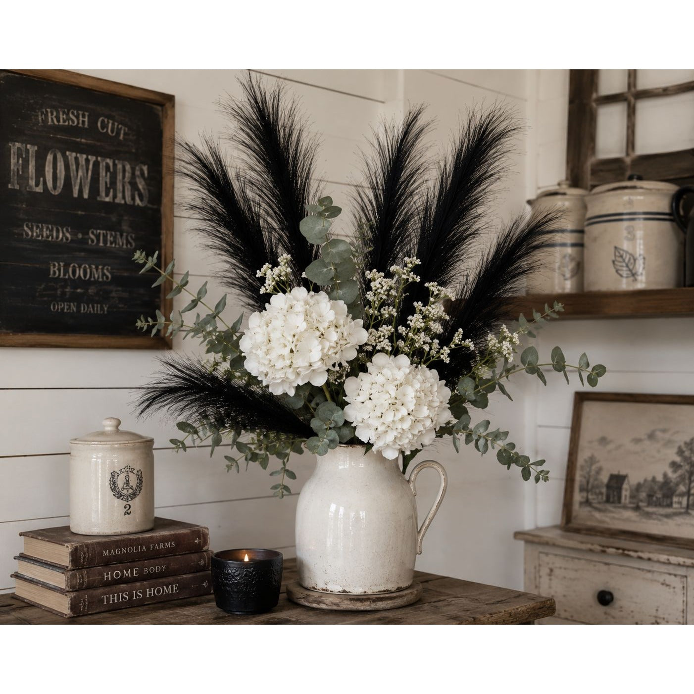 Floral arrangement in a white pitcher with black feathers on a wooden surface, with a rustic decor background.