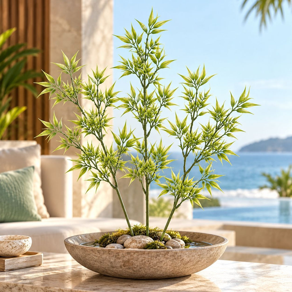 A potted artificial bamboo plant with green and yellow variegated leaves in a shallow stone bowl filled with rocks and moss, set on a marble table with a blurred outdoor patio and ocean background.