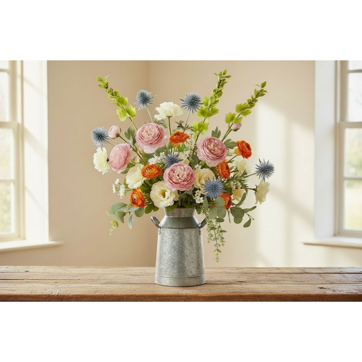 A floral arrangement of pink, orange, white, and blue flowers in a galvanized metal milk canister vase, displayed on a rustic wooden table in front of a softly lit beige wall and window.