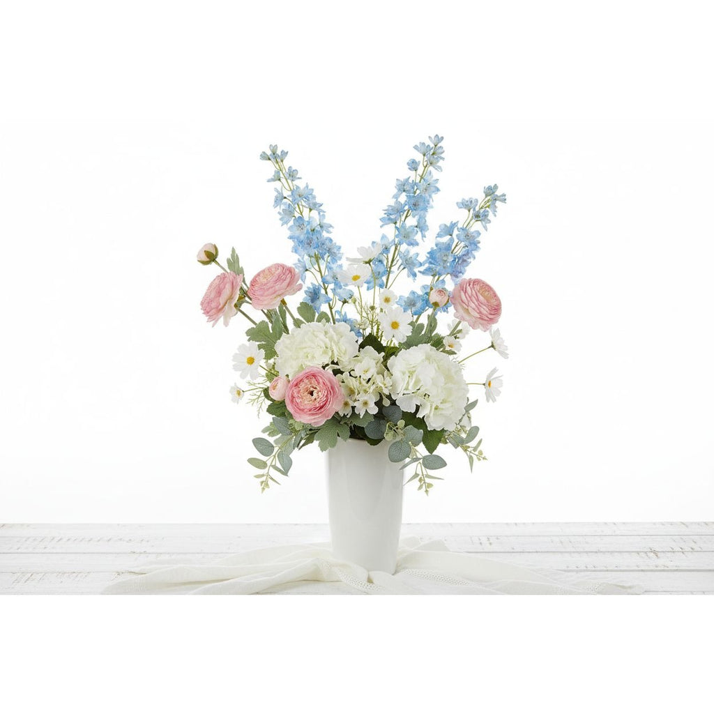 A floral arrangement in a white ceramic vase, featuring pink roses, white ranunculus, light blue delphiniums, and green foliage, set against a clean white background.