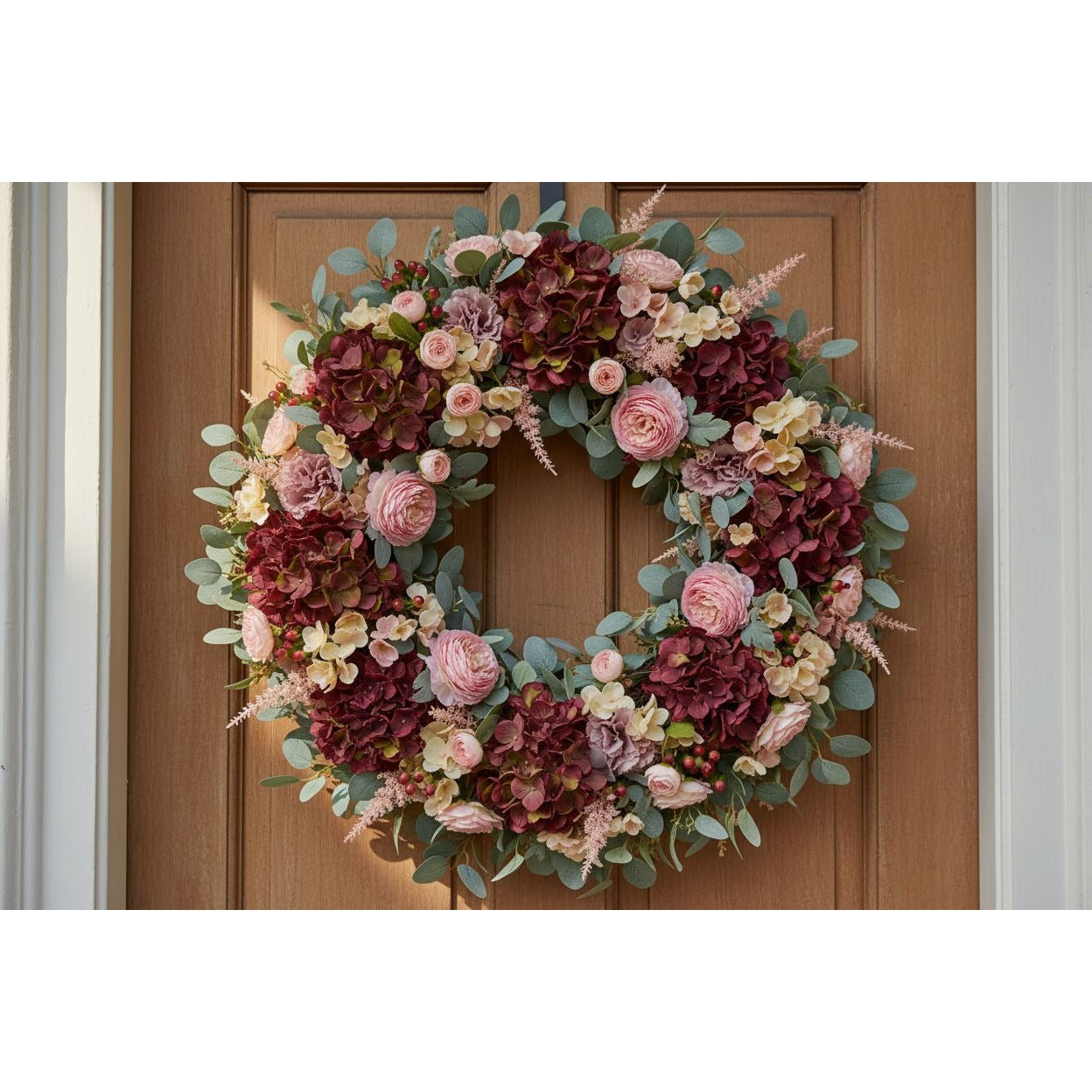 A circular floral wreath with burgundy hydrangeas, pink roses, cream blossoms, and green eucalyptus leaves, hanging on a brown wooden front door.