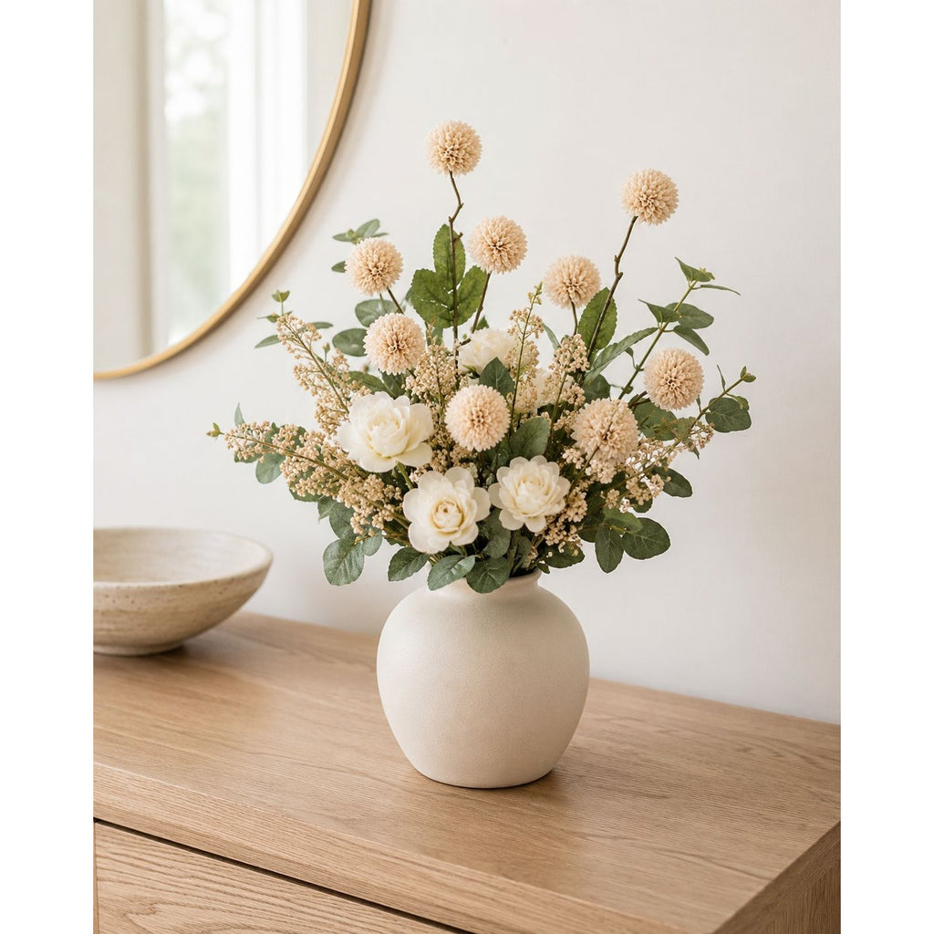 A faux floral arrangement in a matte cream ceramic vase, styled on a light wood console table with a neutral wall and a gold-framed mirror in the background.