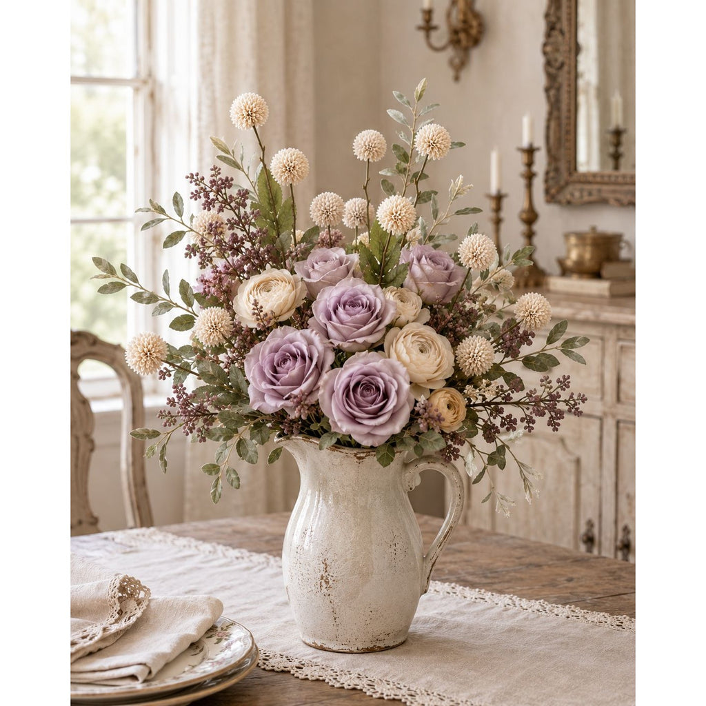 A floral arrangement of lavender and cream roses with green foliage in a distressed white ceramic pitcher, set on a table in a softly lit, vintage-style room.