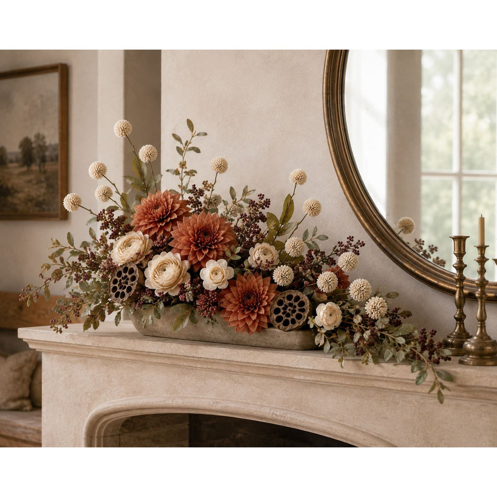 A decorative floral arrangement in a long rectangular planter sits on a fireplace mantel in a styled interior. The arrangement includes rust-orange and cream flowers, green foliage, and dark berry-like accents.