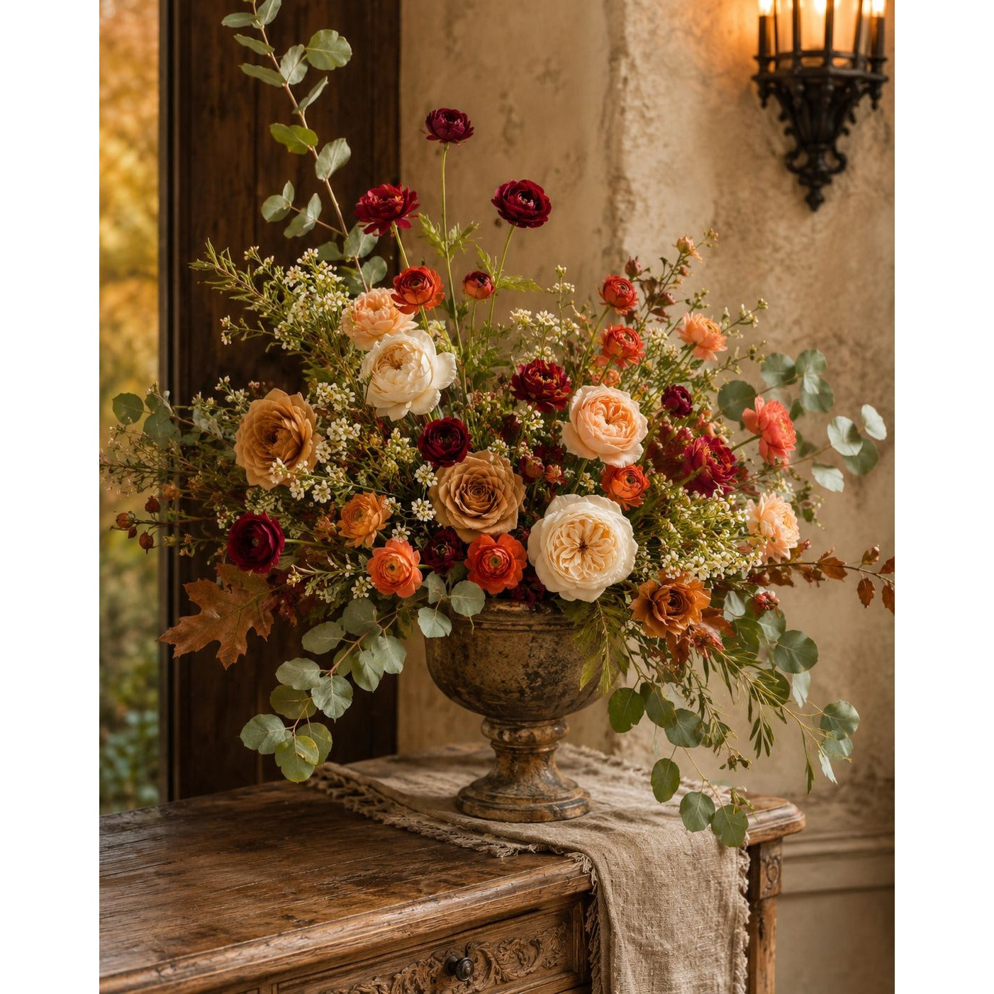 A large, rustic floral arrangement in an antique-style bronze urn, placed on a wooden console table in a warm, vintage room. The bouquet features a mix of orange, red, cream, and burgundy flowers with green eucalyptus foliage.