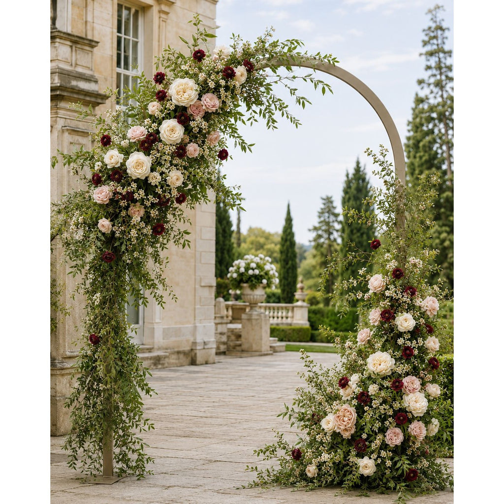 A large arched floral gateway decorated with green vines, white and blush pink flowers, and deep burgundy blooms, set outdoors in front of a stone building and garden trees.