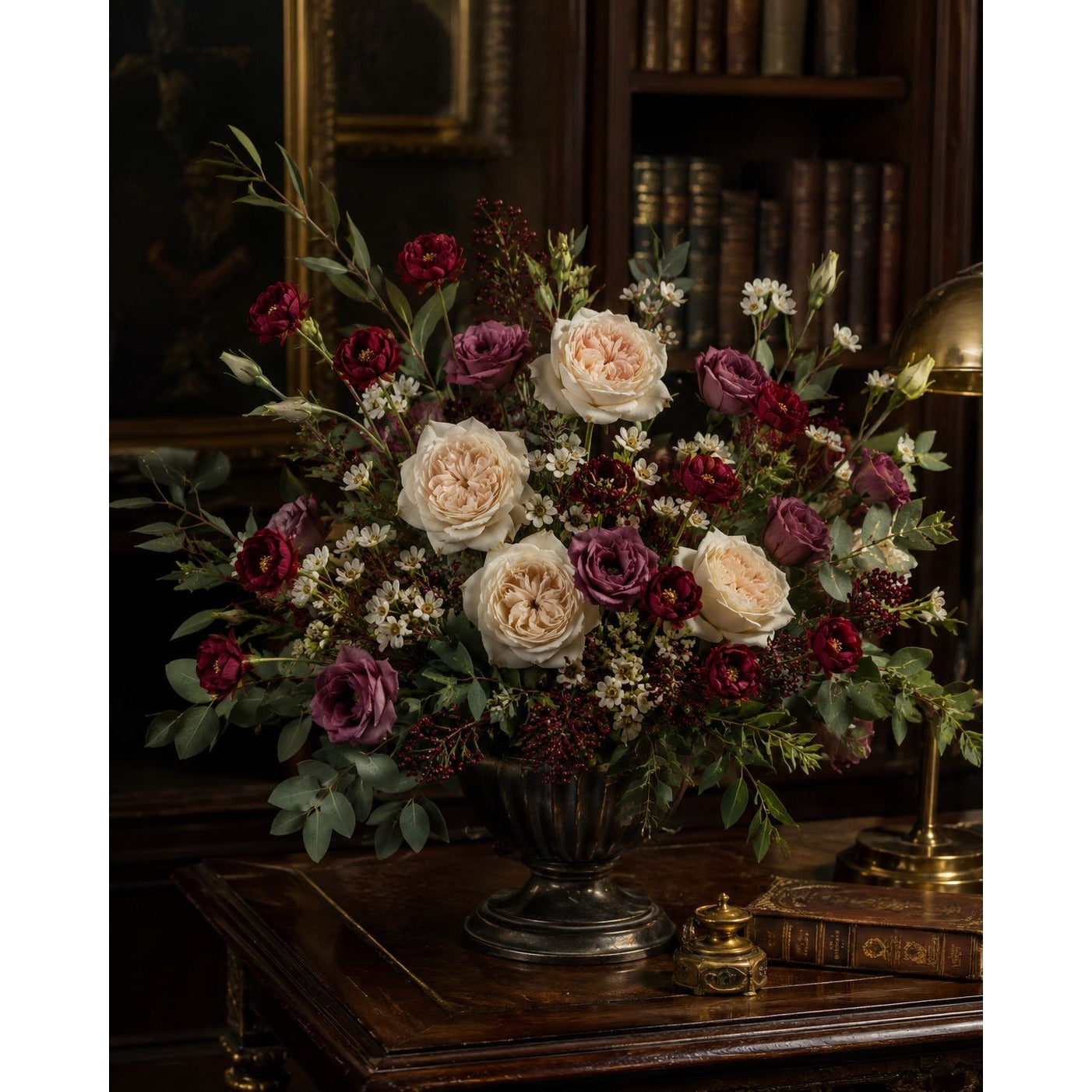 A large floral arrangement in an ornate dark metal urn, featuring cream roses, burgundy roses, mauve-purple roses, and green foliage, displayed on a dark wooden table in a vintage library setting.