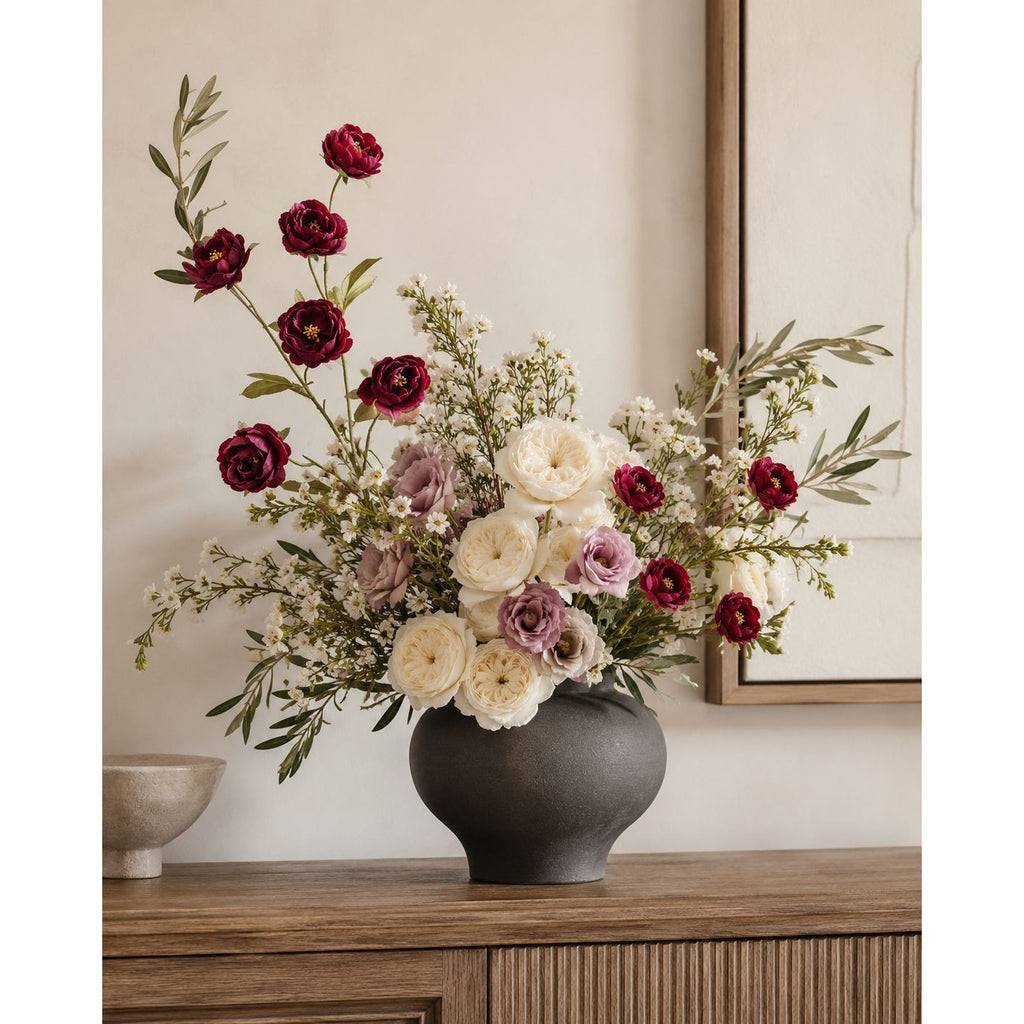 A floral arrangement in a matte dark gray vase, placed on a wooden console table against a warm beige wall. The bouquet features deep burgundy roses, cream ranunculus, soft lavender flowers, and green foliage.