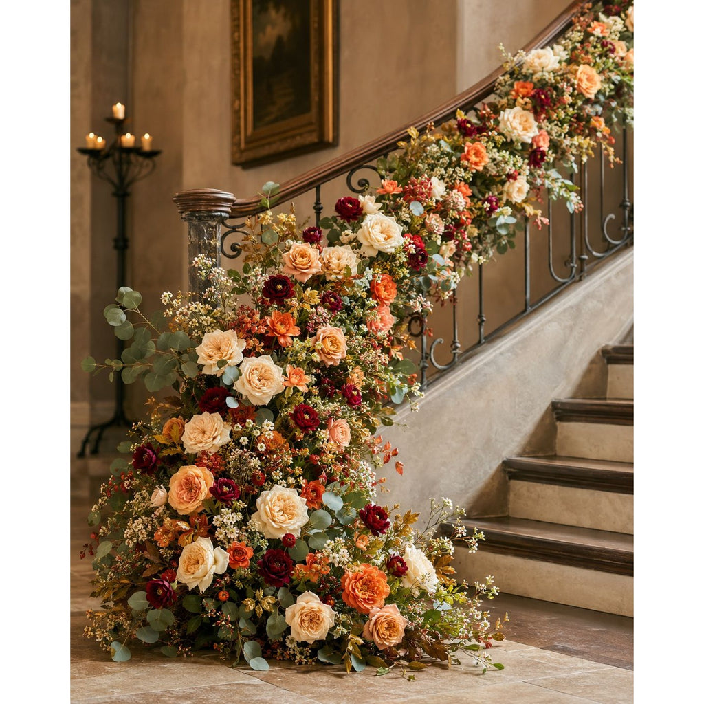 A large floral arrangement cascading down the base of an indoor staircase, with additional flowers draped along the curved banister. The arrangement features cream, peach, orange, burgundy, and burgundy roses with green foliage, set against a warm beige interior wall and stone steps.