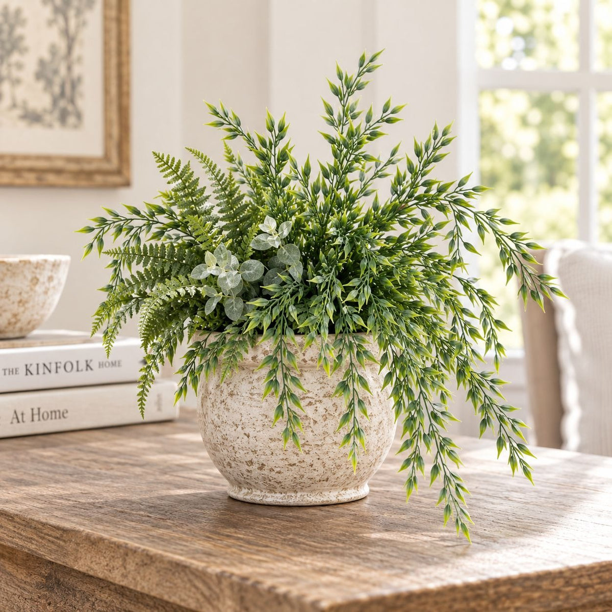An artificial green fern plant in a speckled beige ceramic pot, styled on a wooden table in a bright, modern living room setting.