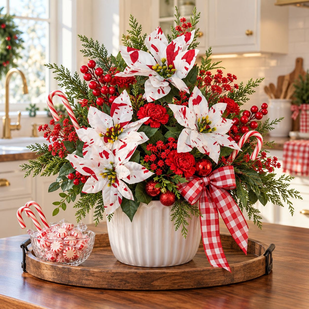 A festive Christmas floral arrangement in a white ribbed pot, featuring white poinsettias with red spots, red carnations, green pine branches, red berries, candy canes, and a red-and-white gingham ribbon, displayed on a wooden tray in a bright kitchen.