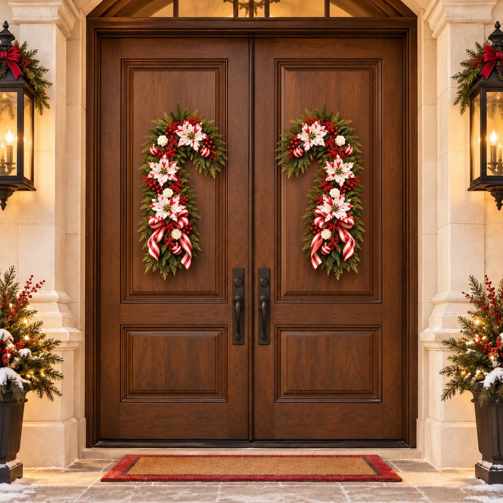 A pair of dark brown wooden front doors decorated with two matching Christmas wreaths featuring red and white ribbons, white flowers, and green foliage. The doors are set in a stone entryway with lit wall lanterns, potted holiday plants, and a red-bordered doormat.
