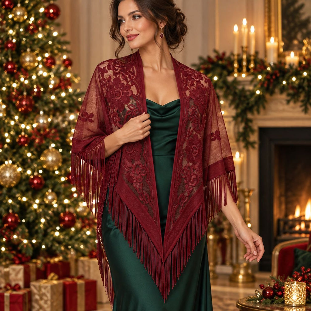 A woman stands in a decorated holiday room wearing a burgundy lace and fringe shawl over a dark green satin dress. A lit Christmas tree, fireplace, candles, and wrapped gifts are visible in the background.