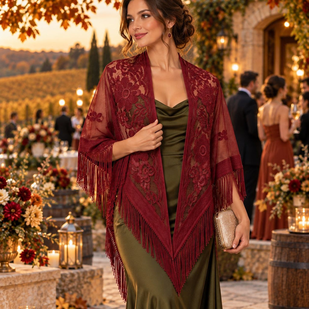 A woman at an outdoor evening event wears a burgundy lace and fringe shawl over an olive green dress, holding a small metallic clutch. The background shows a vineyard and warmly lit guests.