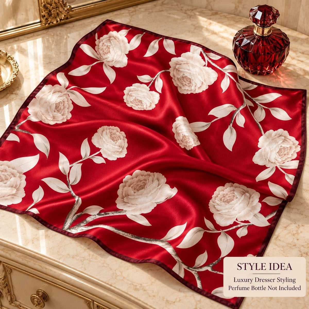 A red satin scarf with a large white rose and leaf pattern, styled on a marble vanity next to a red perfume bottle. A text overlay in the bottom right corner reads 'STYLE IDEA Luxury Dresser Styling Perfume Bottle Not Included'.