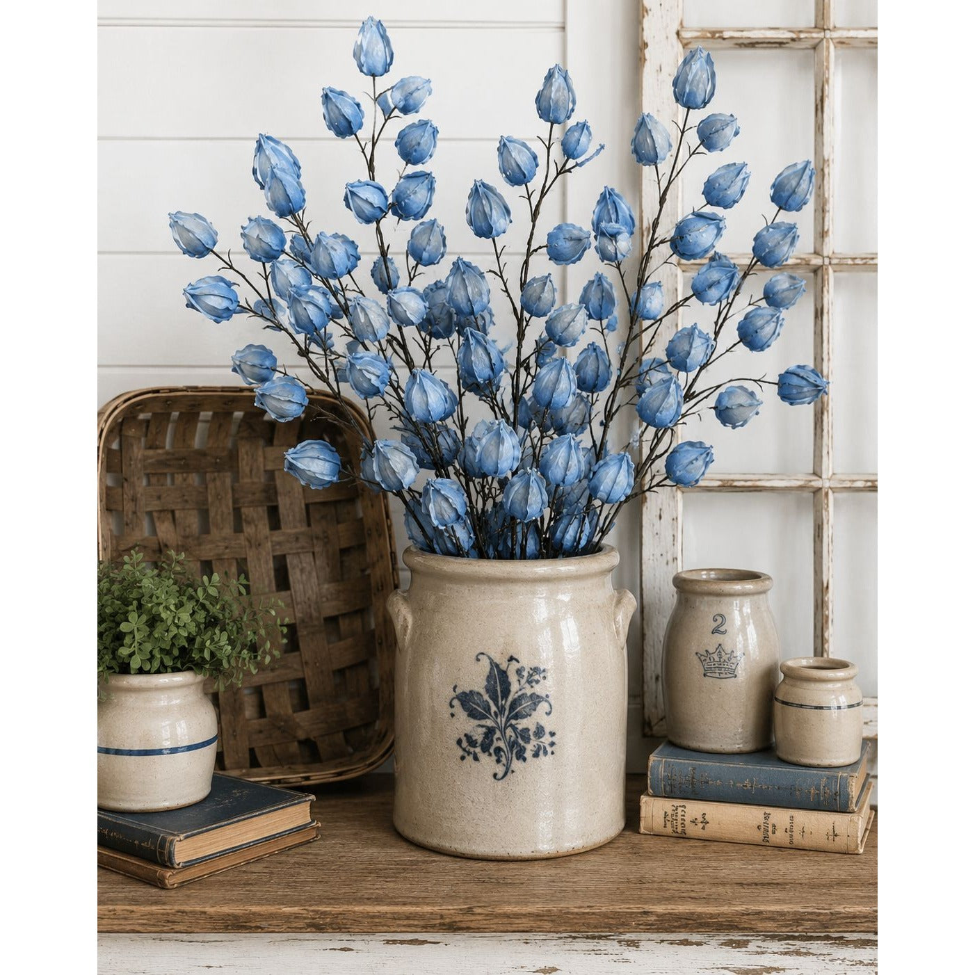 Decorative blue floral arrangement in a rustic ceramic vase on a wooden surface with books and a basket.