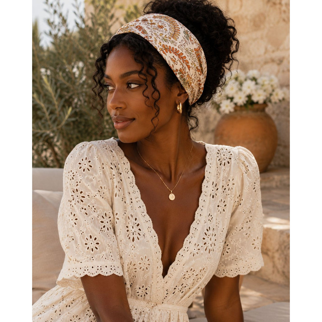 A woman wearing a cream-colored eyelet lace dress with puff sleeves and a matching headband, posed in an outdoor setting with a stone wall and potted flowers in the background.