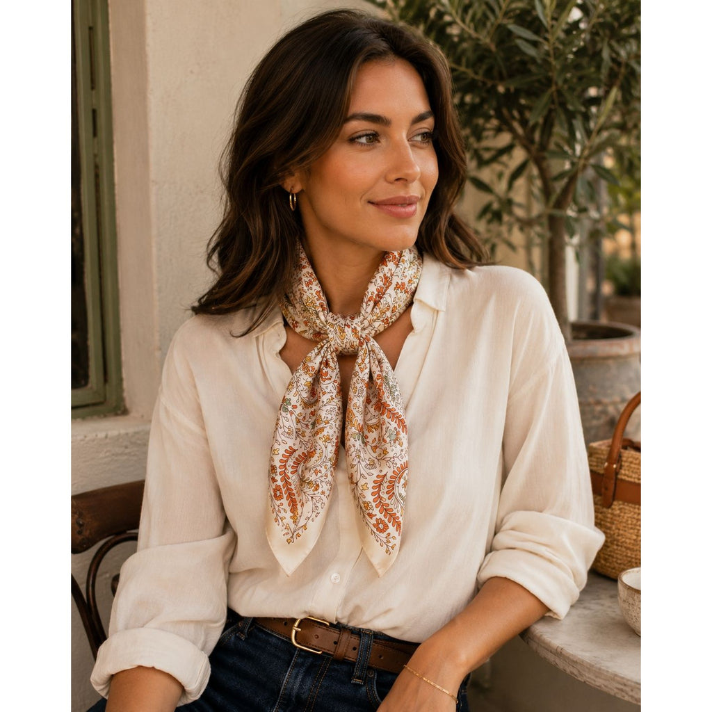 A woman wearing a cream-colored button-up blouse and a patterned scarf with rust-orange and beige paisley details, sitting in an outdoor patio setting.