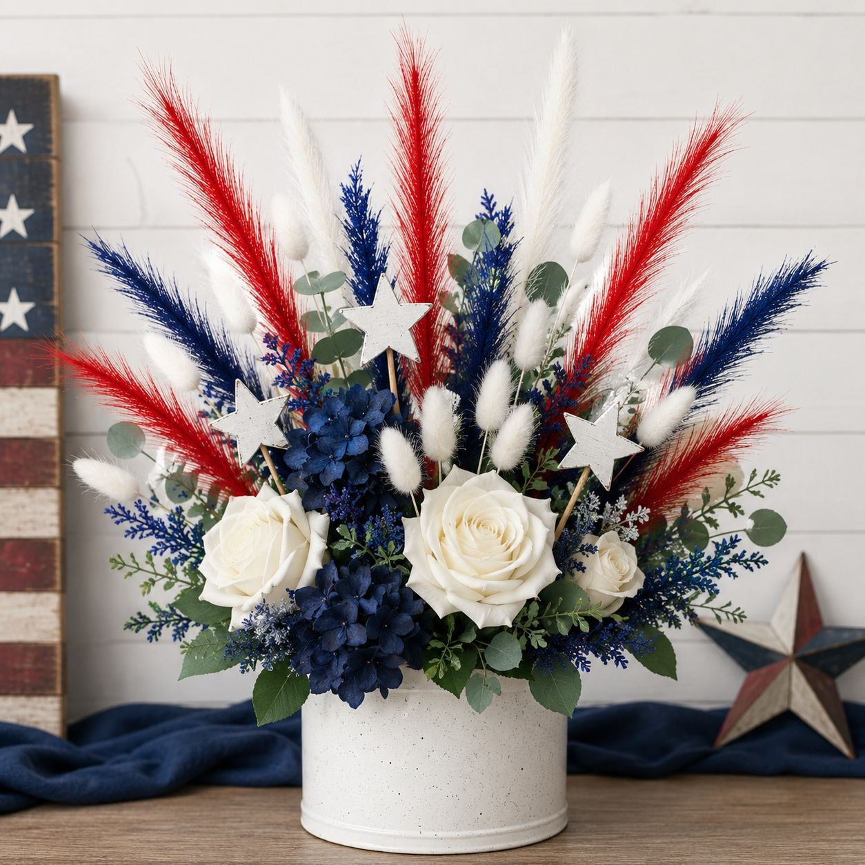 Patriotic floral arrangement in a white speckled bucket vase, featuring white roses, blue hydrangeas, red pampas grass, white and blue bunny tails, eucalyptus leaves, and white star cutouts, displayed on a wooden surface against a white shiplap wall.