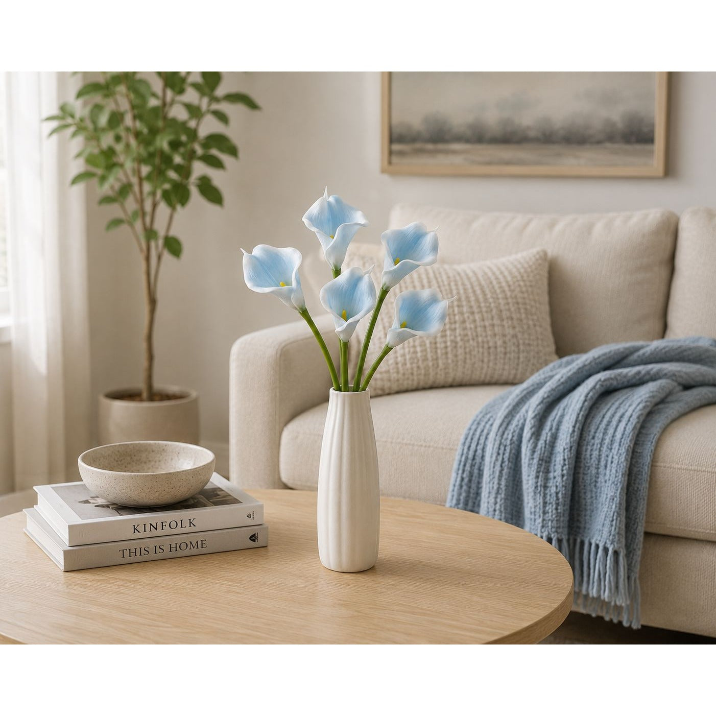 A tall ribbed off-white ceramic vase holding light blue calla lilies, placed on a round light wood coffee table in a softly styled living room with a cream sofa, blue throw blanket, books, and a potted plant.