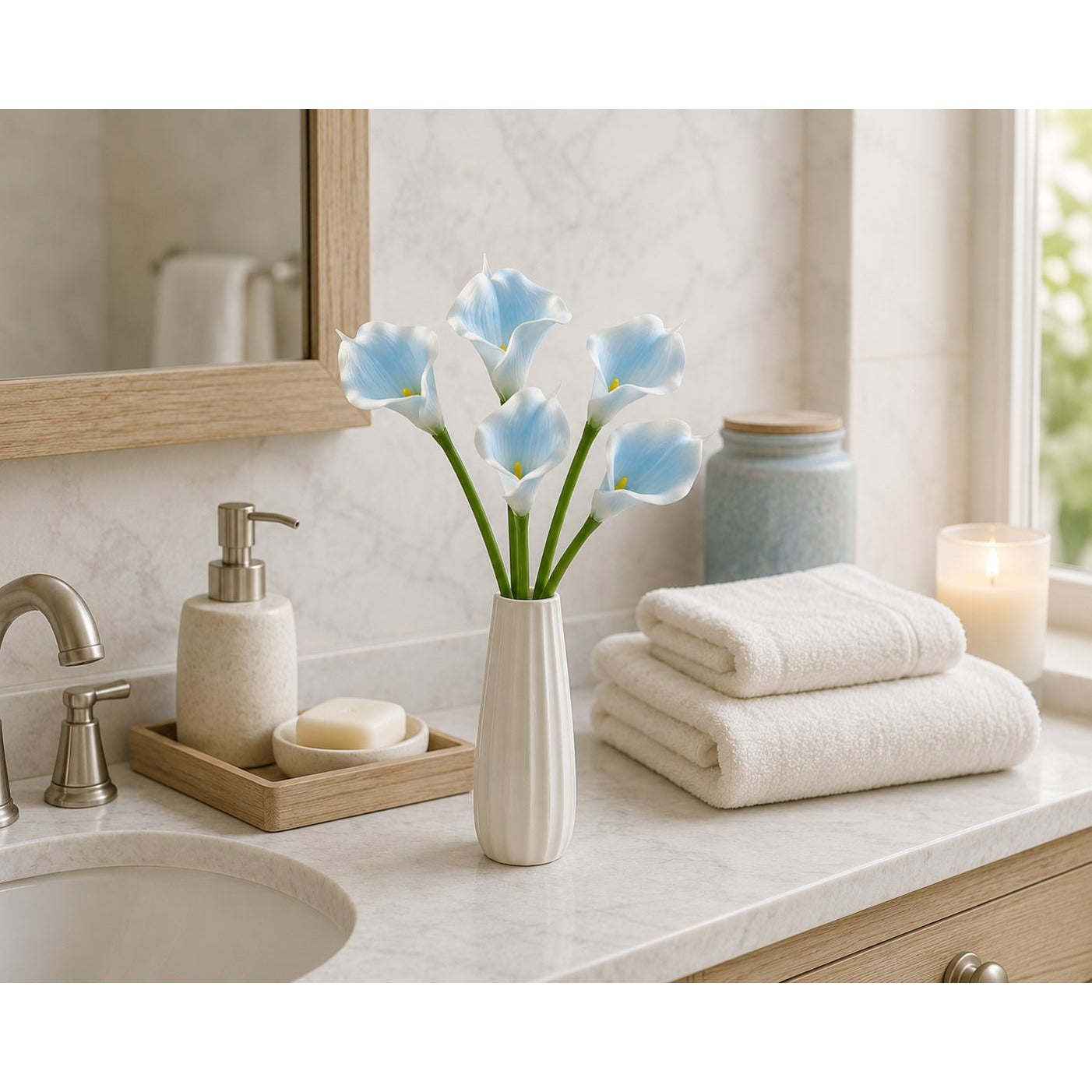 A styled bathroom countertop scene with folded white towels, a vase of light blue calla lilies, a soap dispenser, a tray with soap, and a lit candle near a window.