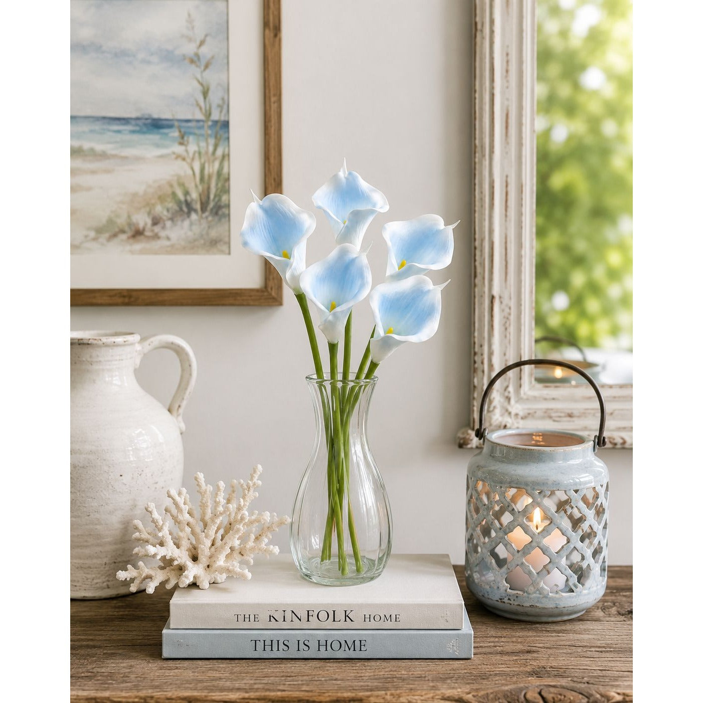 A bouquet of light blue calla lilies in a clear glass vase, styled on a wooden surface with books, a ceramic pitcher, a coral shell, and a lit decorative lantern in a bright indoor setting.