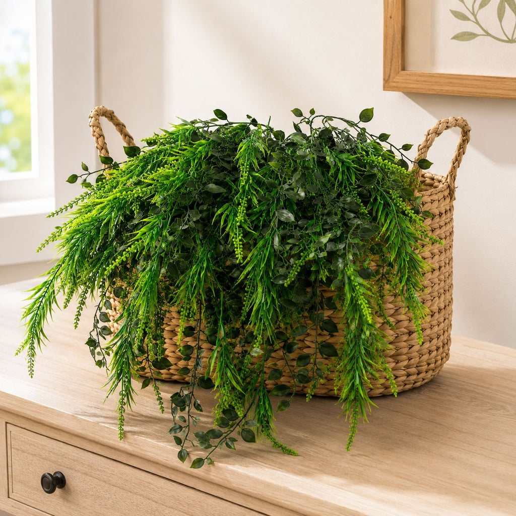 A natural woven seagrass basket with two handles, filled with lush green artificial foliage, displayed on a light wood dresser in a bright room.