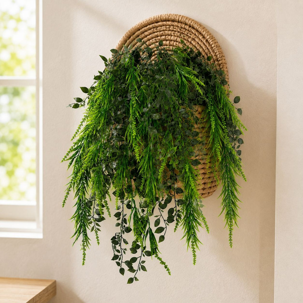 A green artificial trailing plant with dense foliage hanging from a round woven wicker basket mounted on a beige wall next to a window.