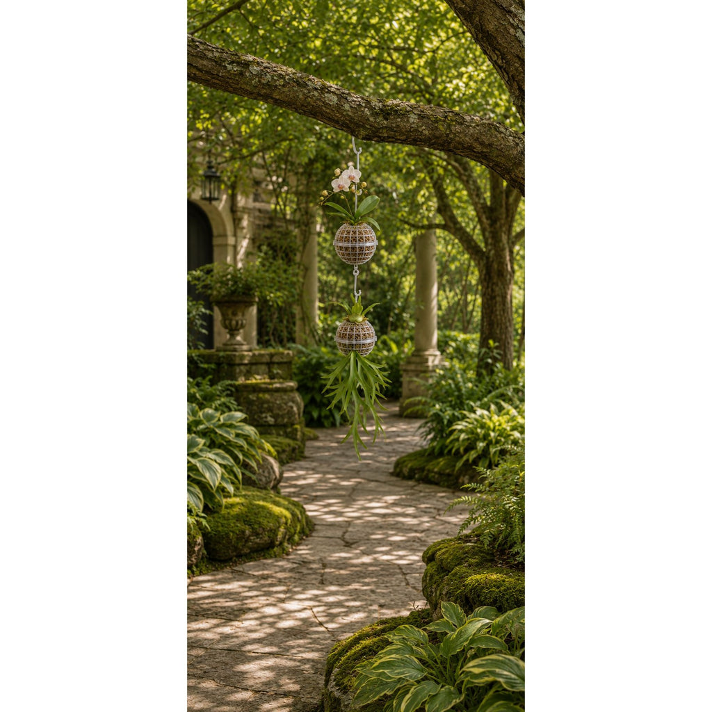 A hanging woven wind spinner with green leaves and small pink flowers suspended from a tree branch in a lush garden pathway.