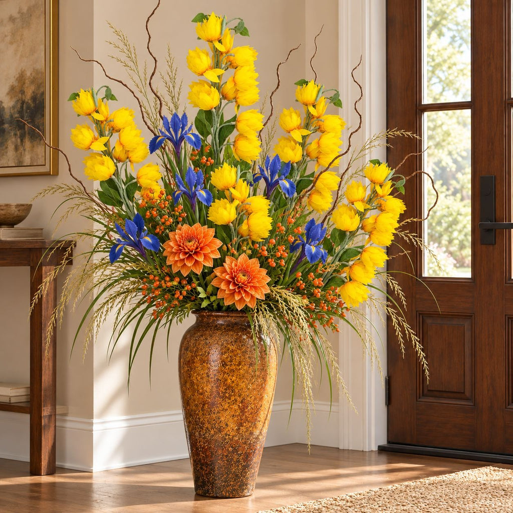 A large artificial floral arrangement in a tall textured brown ceramic vase, placed indoors near a wooden door. The bouquet features bright yellow tulips, blue irises, orange dahlias, and green foliage with tan grasses.