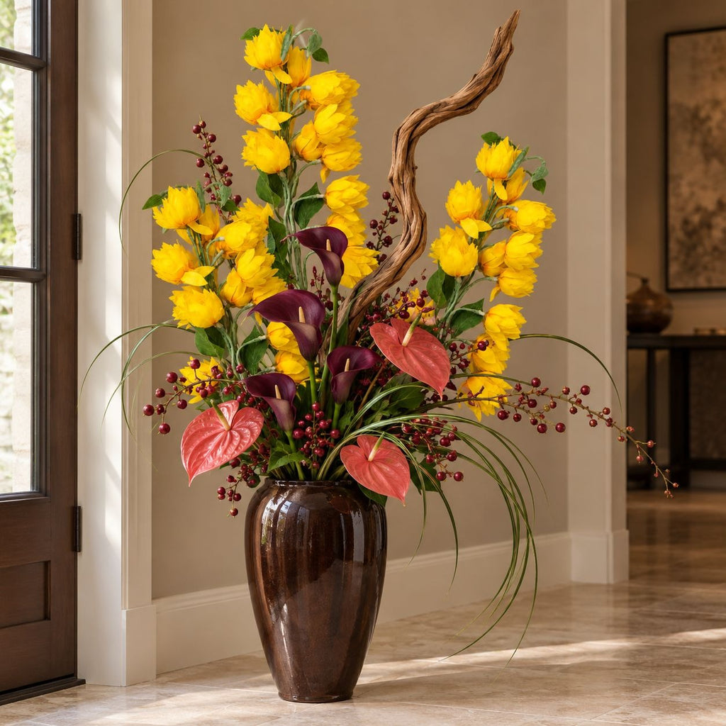 A large artificial floral arrangement in a glossy dark brown vase, featuring bright yellow tulips, dark purple calla lilies, pinkish-red anthuriums, green foliage, and a curved piece of driftwood, displayed indoors on a tiled floor.