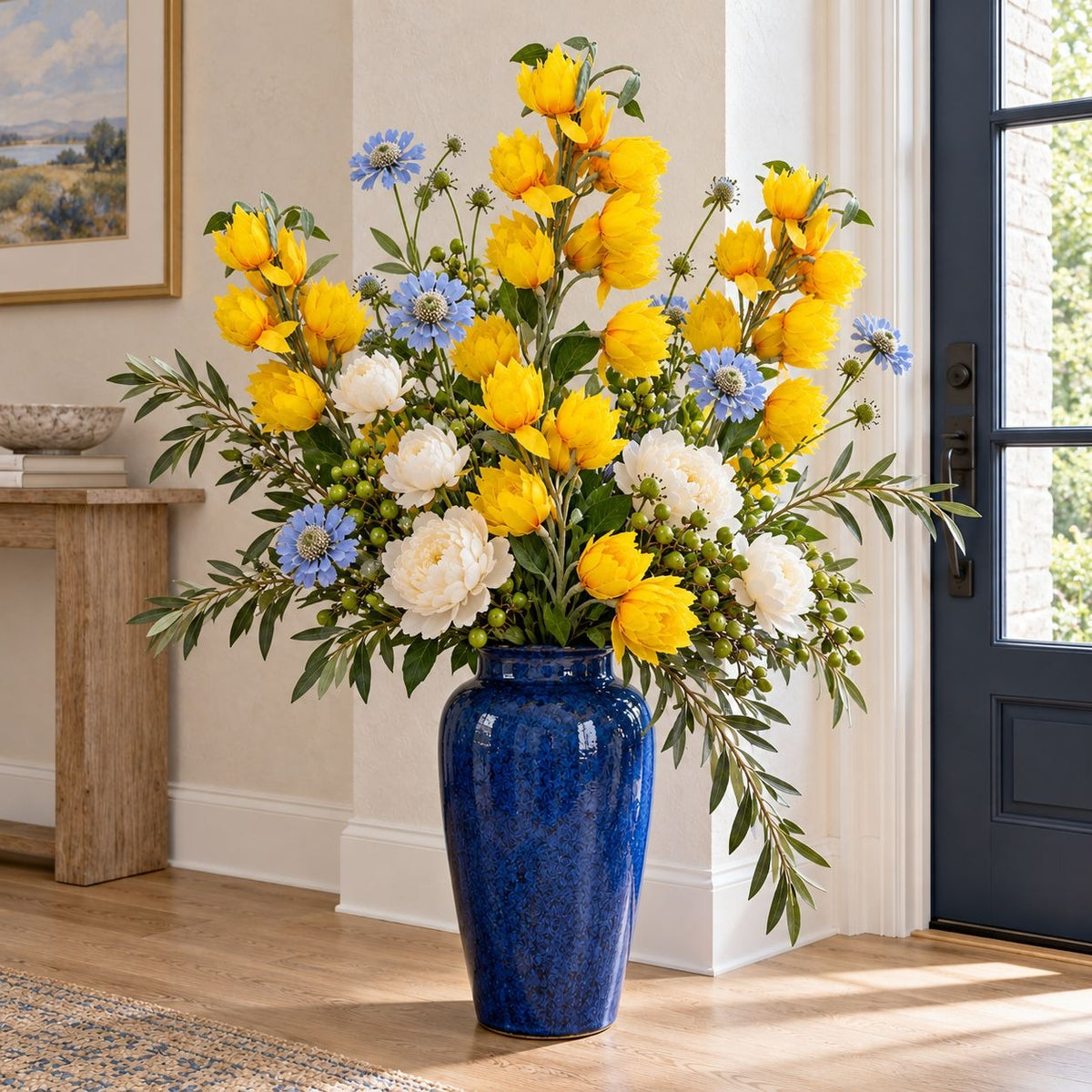 A large blue ceramic vase filled with a vibrant floral arrangement of yellow, white, and blue flowers, placed on a wooden floor in a bright, modern room.