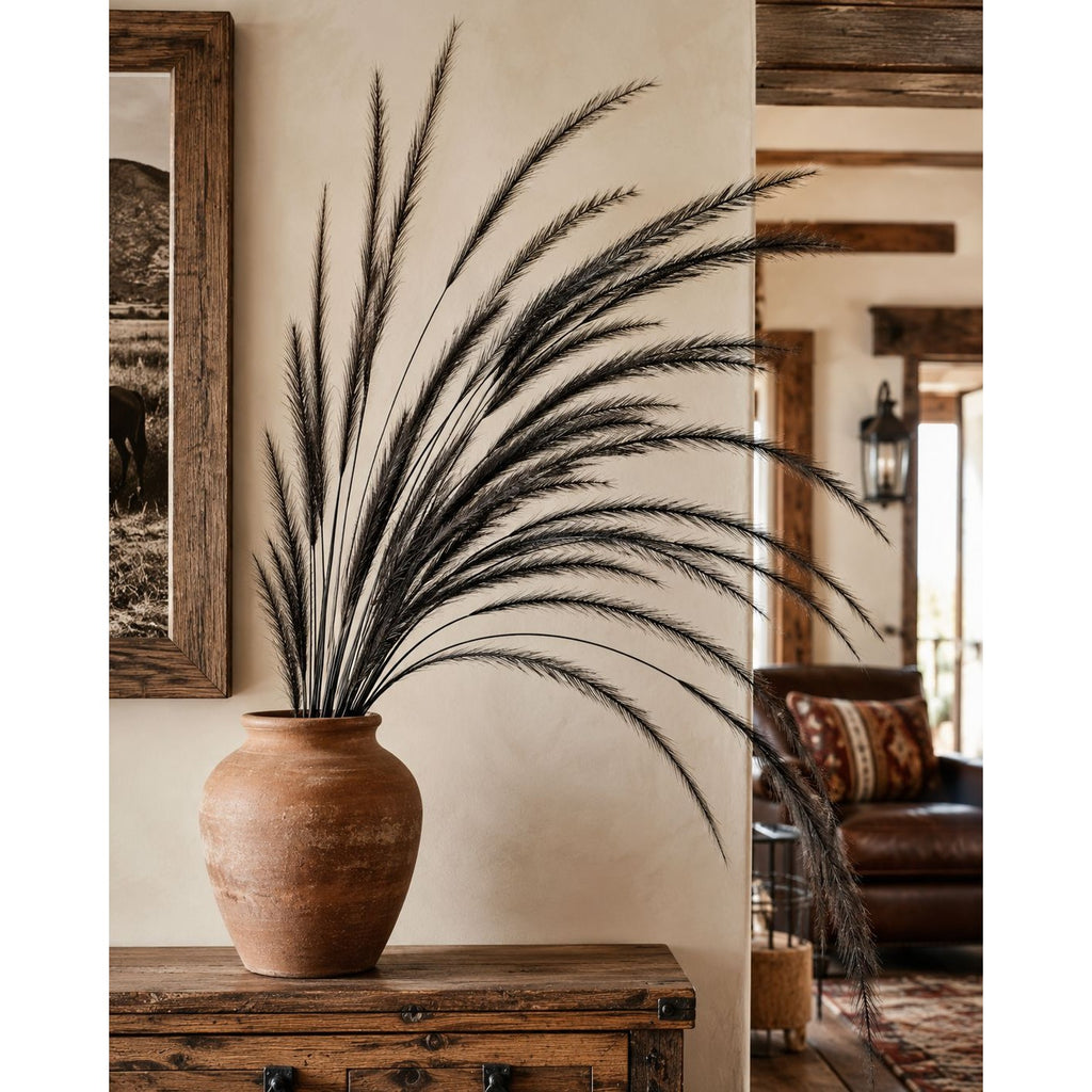 A large terracotta-style ceramic vase filled with tall, wispy dark dried grass stems, displayed on a rustic wooden console table in a warm, rustic interior with a framed print and leather sofa in the background.