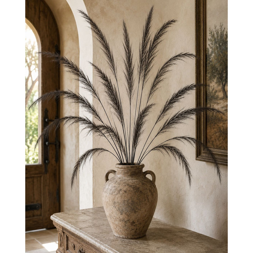 A large rustic beige ceramic vase filled with tall dark pampas grass, displayed on a stone console table in a warm, arched interior room.