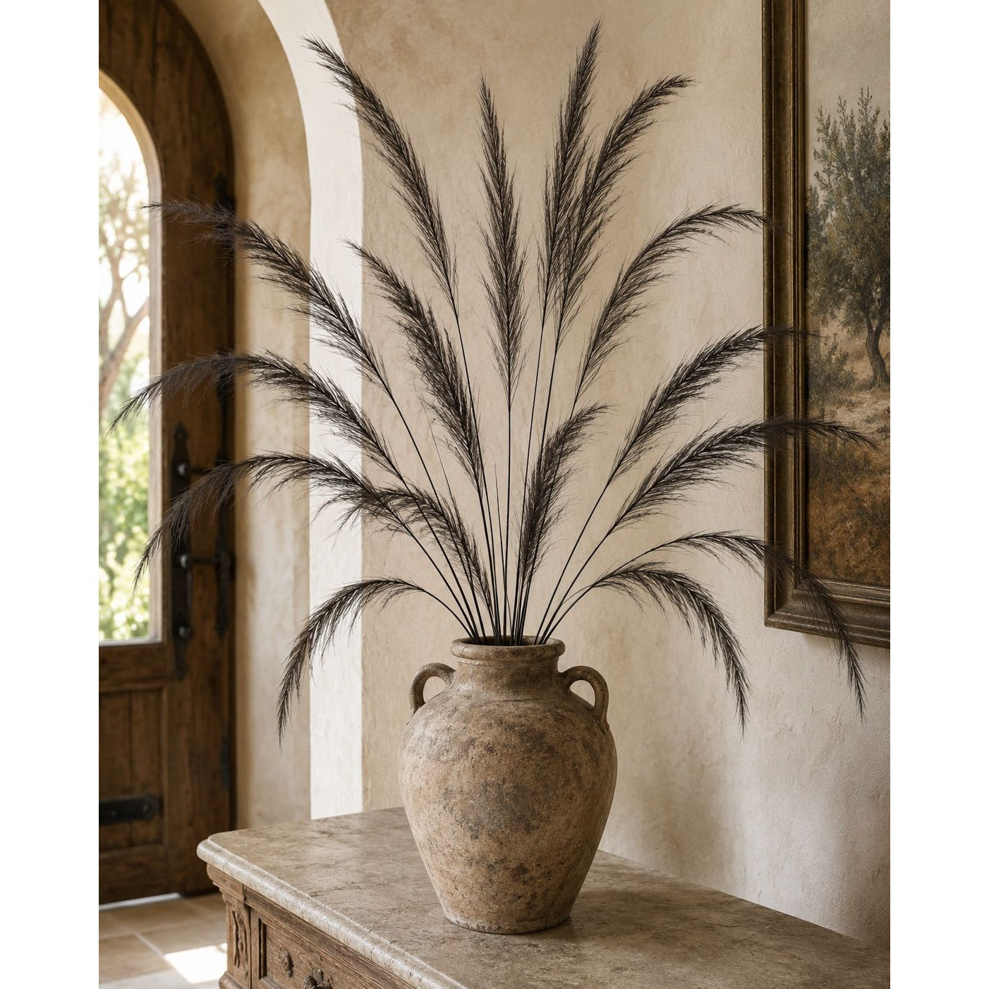 A large rustic beige ceramic vase filled with tall dark pampas grass, displayed on a stone console table in a warm, arched interior room.