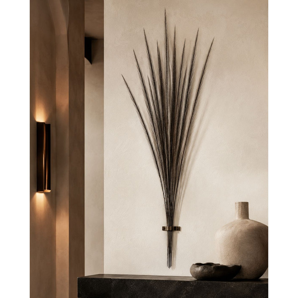 A dark metal wall art piece shaped like a dried palm frond cluster, mounted on a textured beige wall above a dark console table with a ceramic vase and bowl.