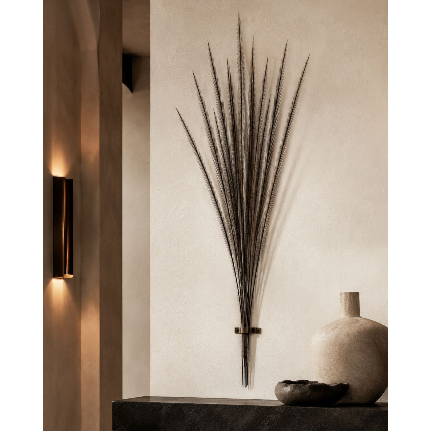 A dark metal wall art piece shaped like a dried palm frond cluster, mounted on a textured beige wall above a dark console table with a ceramic vase and bowl.