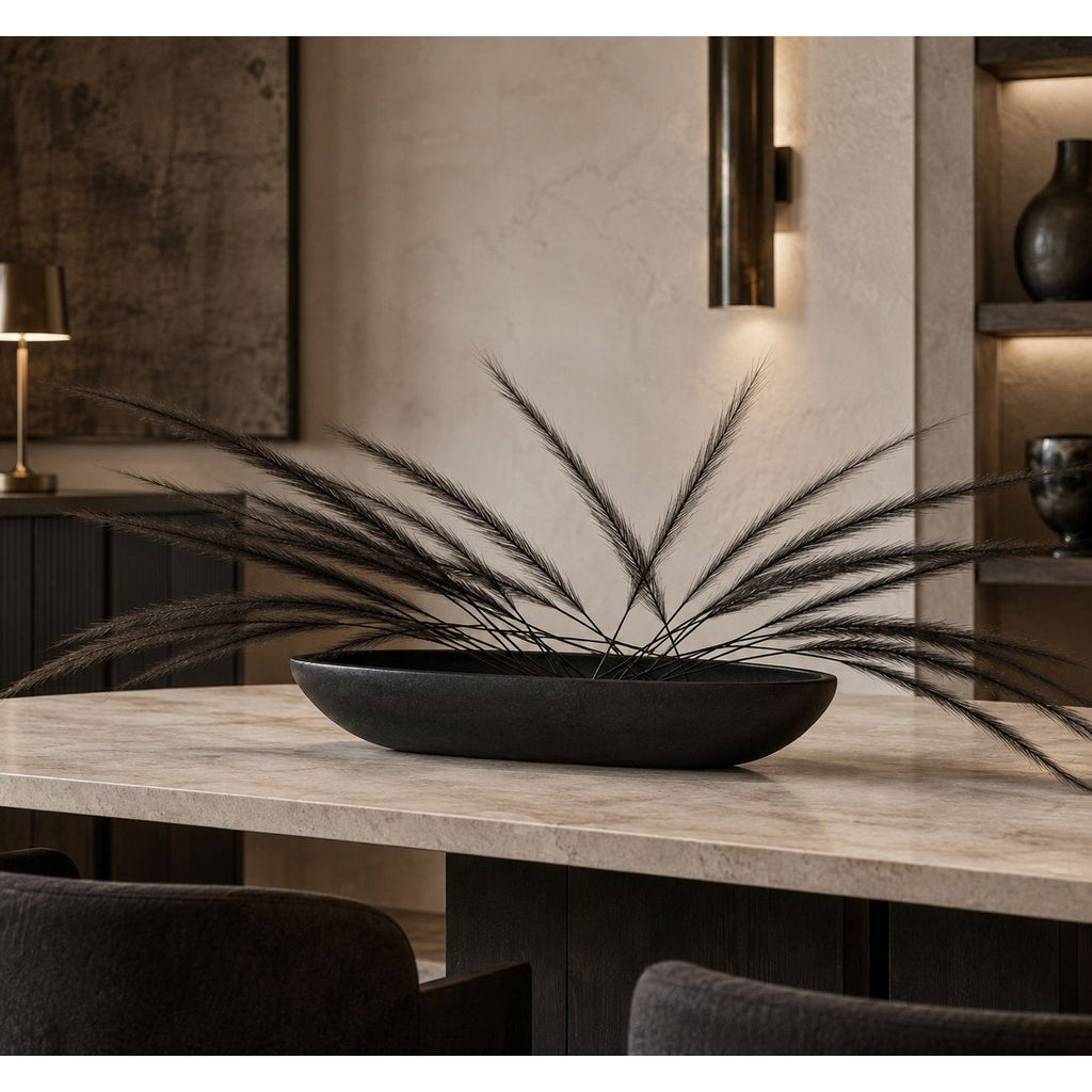 A black oval ceramic planter filled with long, wispy dried grass stems, displayed on a light stone dining table in a modern, dimly lit interior setting.