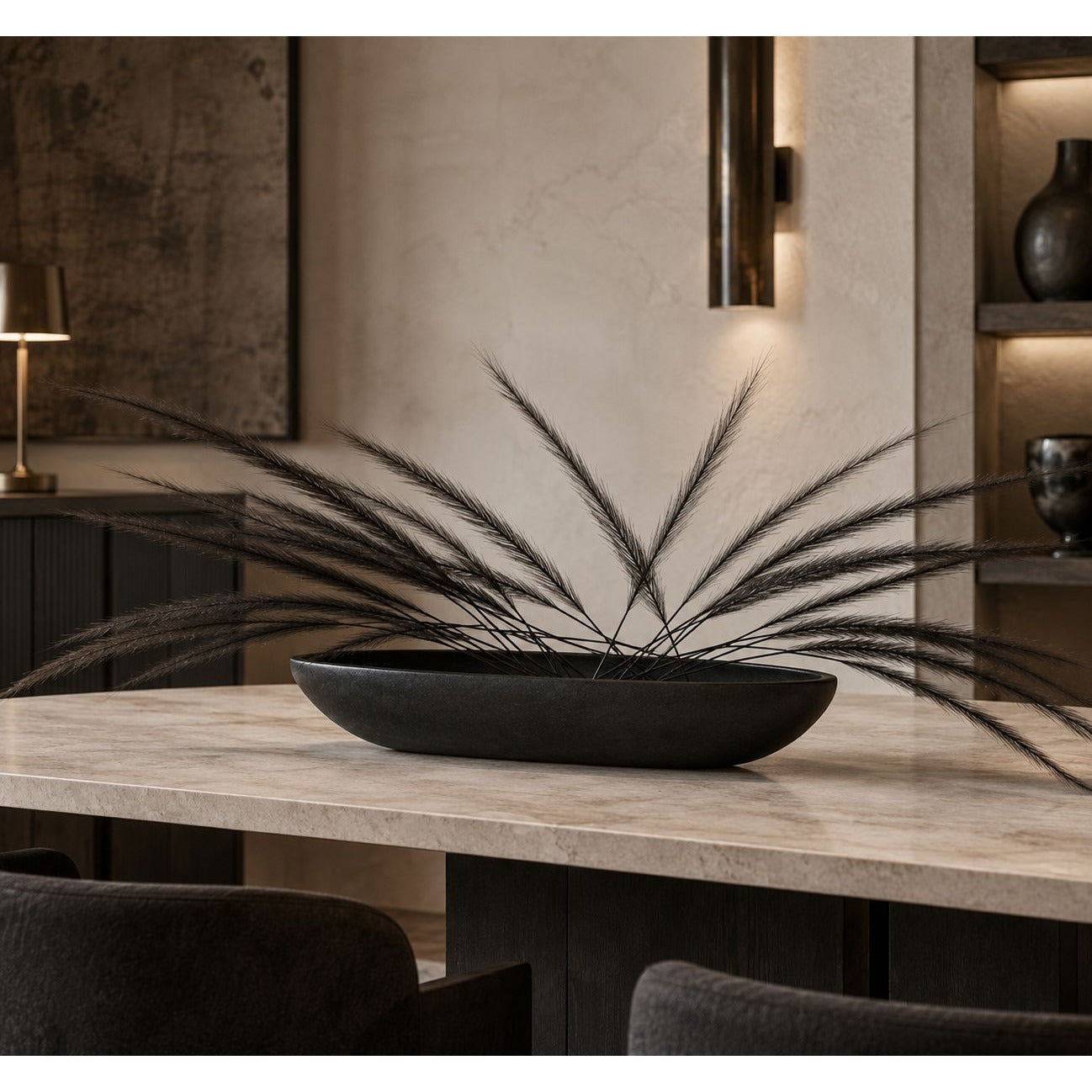 A black oval ceramic planter filled with long, wispy dried grass stems, displayed on a light stone dining table in a modern, dimly lit interior setting.
