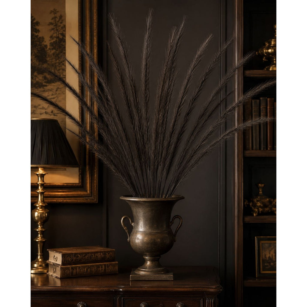 A large bronze-toned urn-shaped vase filled with tall dark dried grass stems, displayed on a dark wooden cabinet in a dimly lit room with a lamp, framed artwork, and bookshelves in the background.