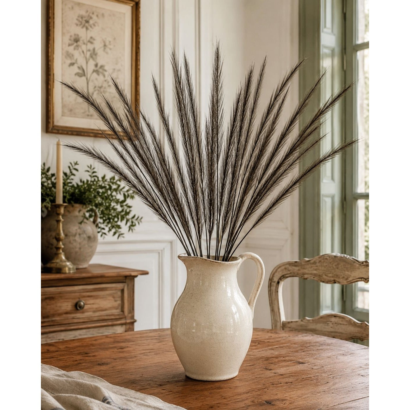 A cream-colored ceramic pitcher vase filled with tall, dark brown ornamental grass stems, set on a rustic wooden table in a bright, traditional room.