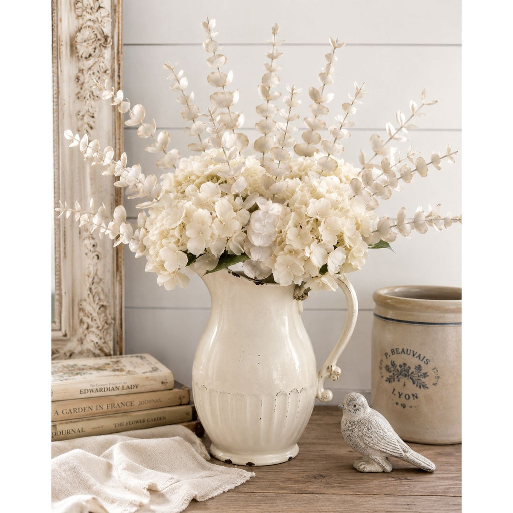 White floral arrangement in a decorative vase on a wooden surface with a mirror and books in the background.
