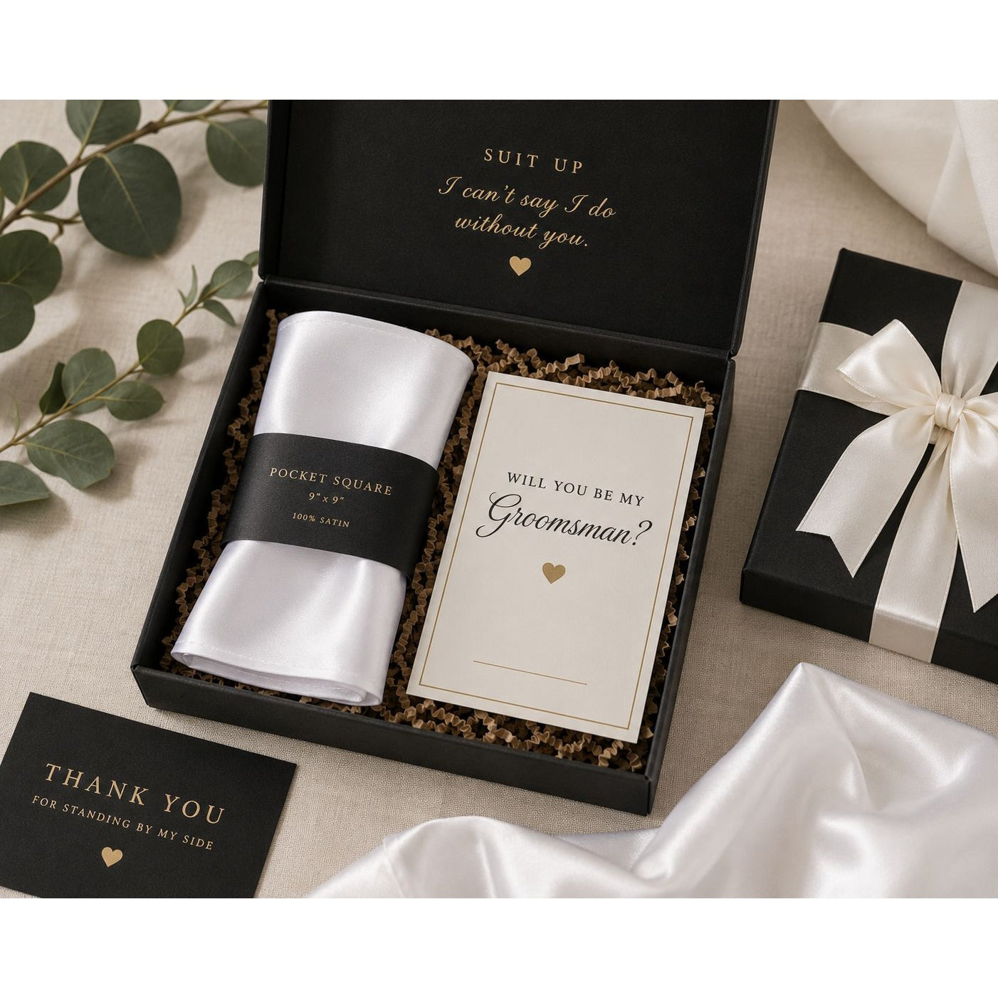 Groomsmen gift set in a black box with a white pocket square, a white wedding card reading 'Will you be my Groomsman?', and a thank you card, styled with greenery and satin fabric.