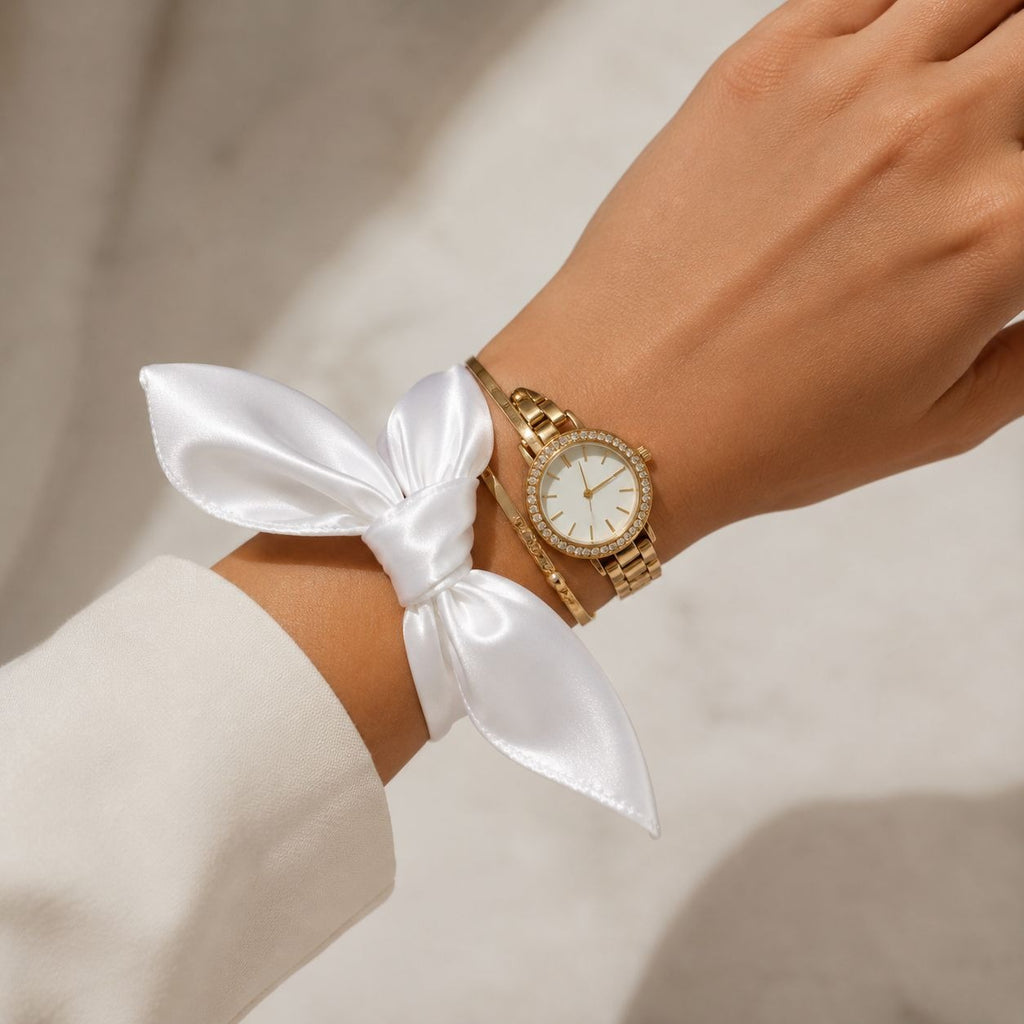 A gold-tone watch with a white dial and crystal-encrusted bezel, worn on a wrist alongside a white satin bow and a gold bracelet, shown on a model's wrist.