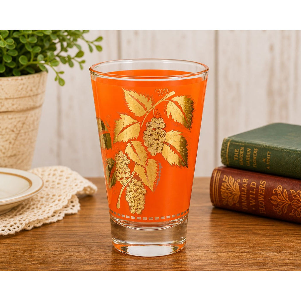 Glass with floral design on an orange background on a wooden table with books and a plant.