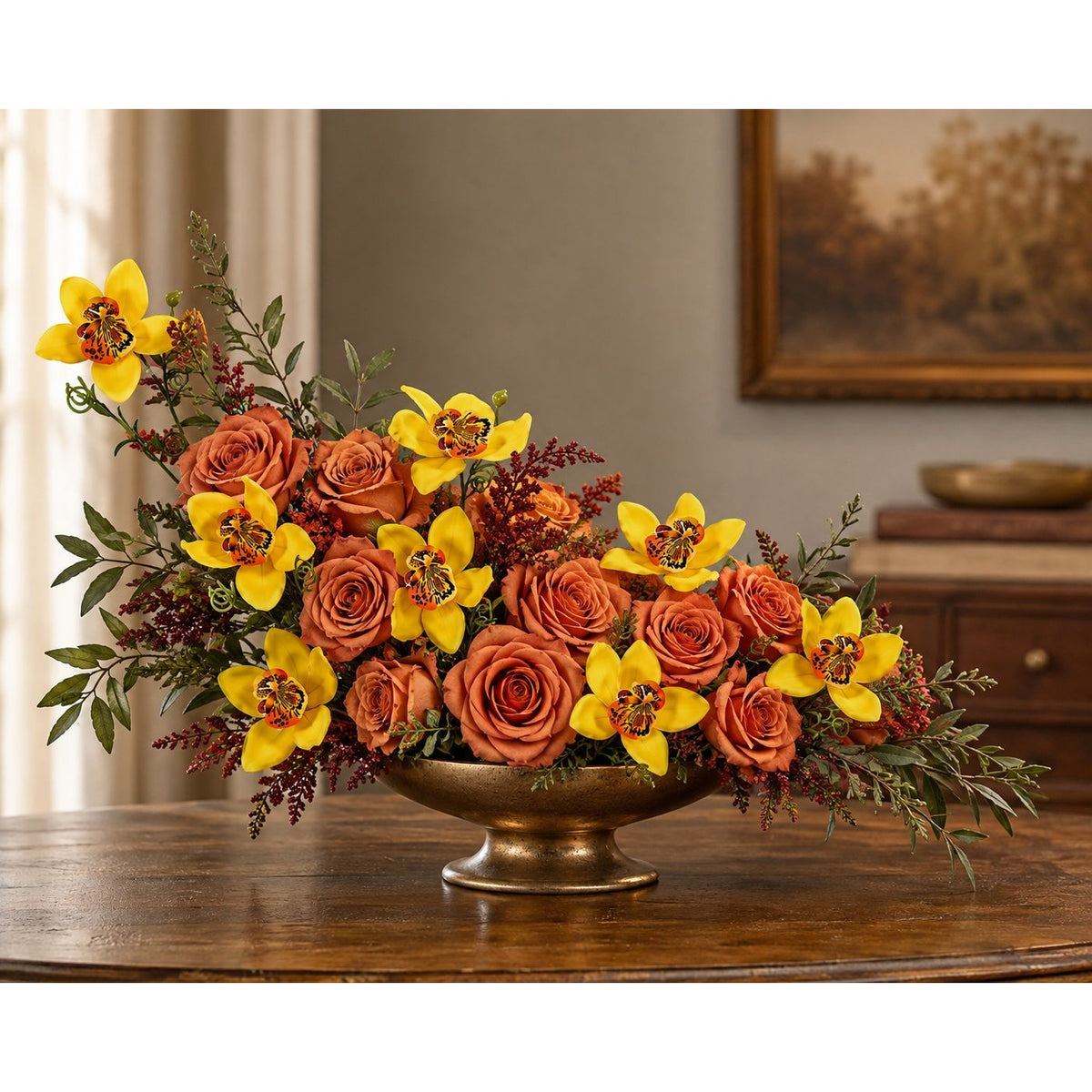 A large autumn floral arrangement featuring orange roses, yellow lilies, and green foliage in a bronze-colored pedestal bowl, displayed on a wooden table in an indoor setting.